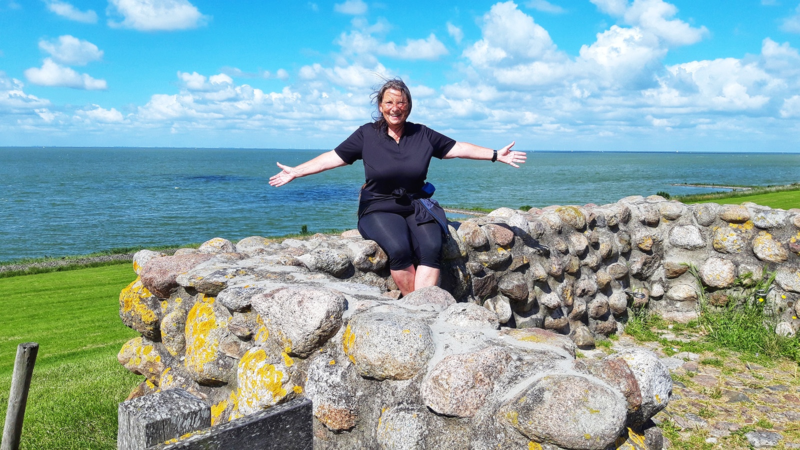 Frau sitzt auf einer Feldsteinmauer am Elfstedenpad, hinter ihr das Ijsselmeer